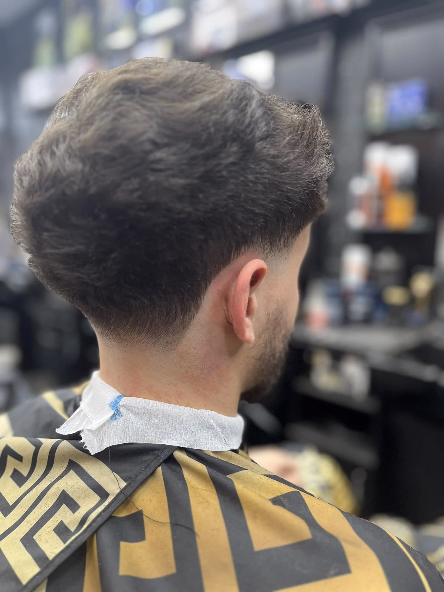 Dense textured hair left long on top over a low taper fade behind the ear, rear three-quarter view